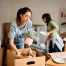 Fatboy Slims Moving Company | Blog Two women unpacking boxes in a living room, placing items like a potted plant and dishes on shelves.