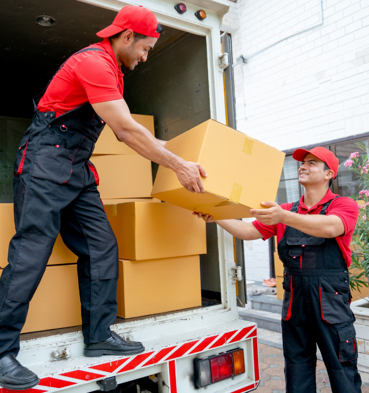 Two workers in red shirts and caps are unloading cardboard boxes from a truck. The scene is outdoors near a brick wall.