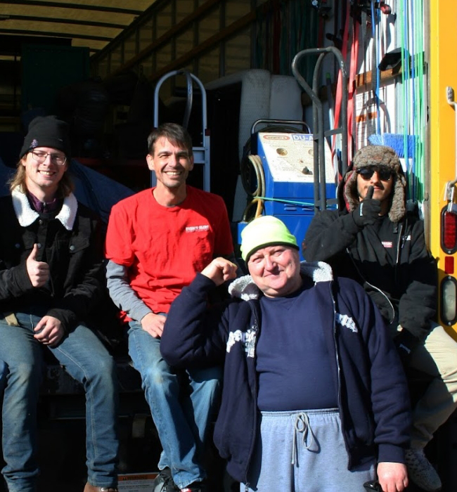 Fatboy Slims Moving Company | Why Us? Four people sitting and standing in a truck, one giving a thumbs up, another gesturing with a peace sign. Bright, casual clothing and a sunny day.