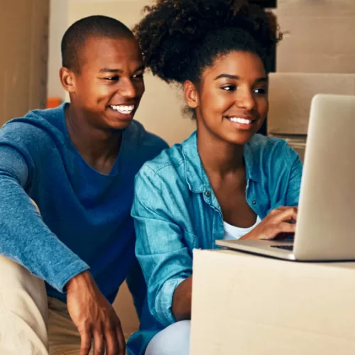 A smiling couple sits on the floor, using a laptop placed on a cardboard box. Both appear engaged with the screen.