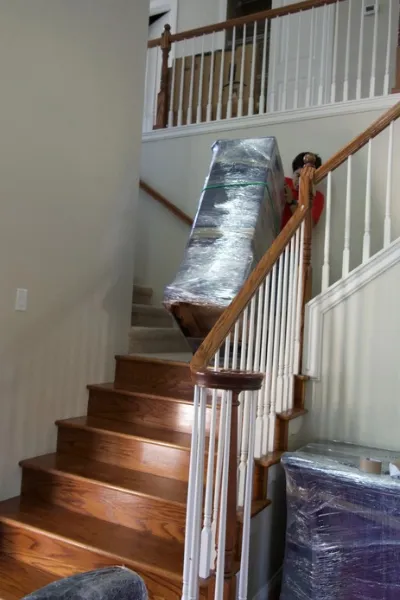Fatboy Slims Moving Company mover carrying wrapped furniture down a staircase.